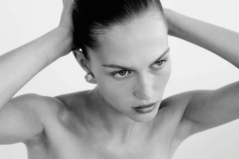 Close-up portrait in black and white of an adult woman with her hands behind her head, looking to the side with a serious expression.
