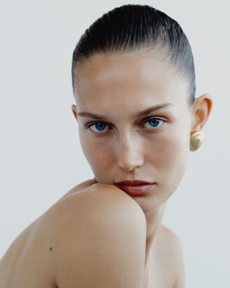 Close-up portrait of a fair-skinned woman with blue eyes and slicked-back hair, wearing gold earrings against a clean, bright background.