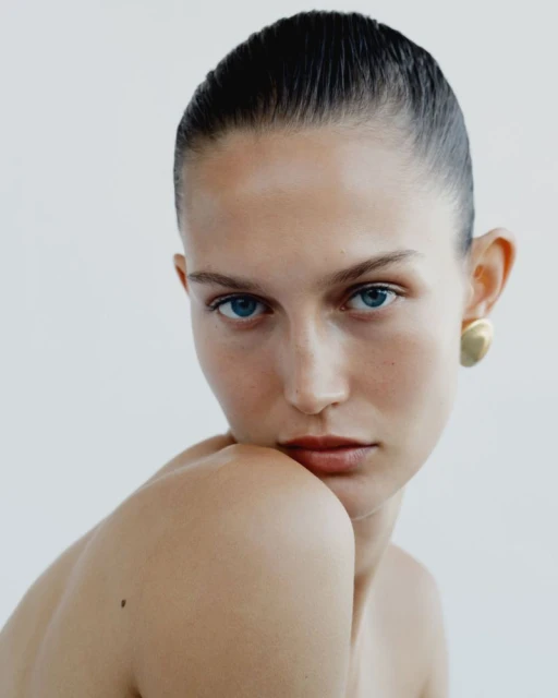 Close-up portrait of a fair-skinned woman with blue eyes and slicked-back hair, wearing gold earrings against a clean, bright background.