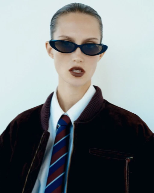 A stylish woman poses in a white shirt, striped tie, and velvet bomber jacket, accessorized with dark sunglasses against a clean white backdrop.