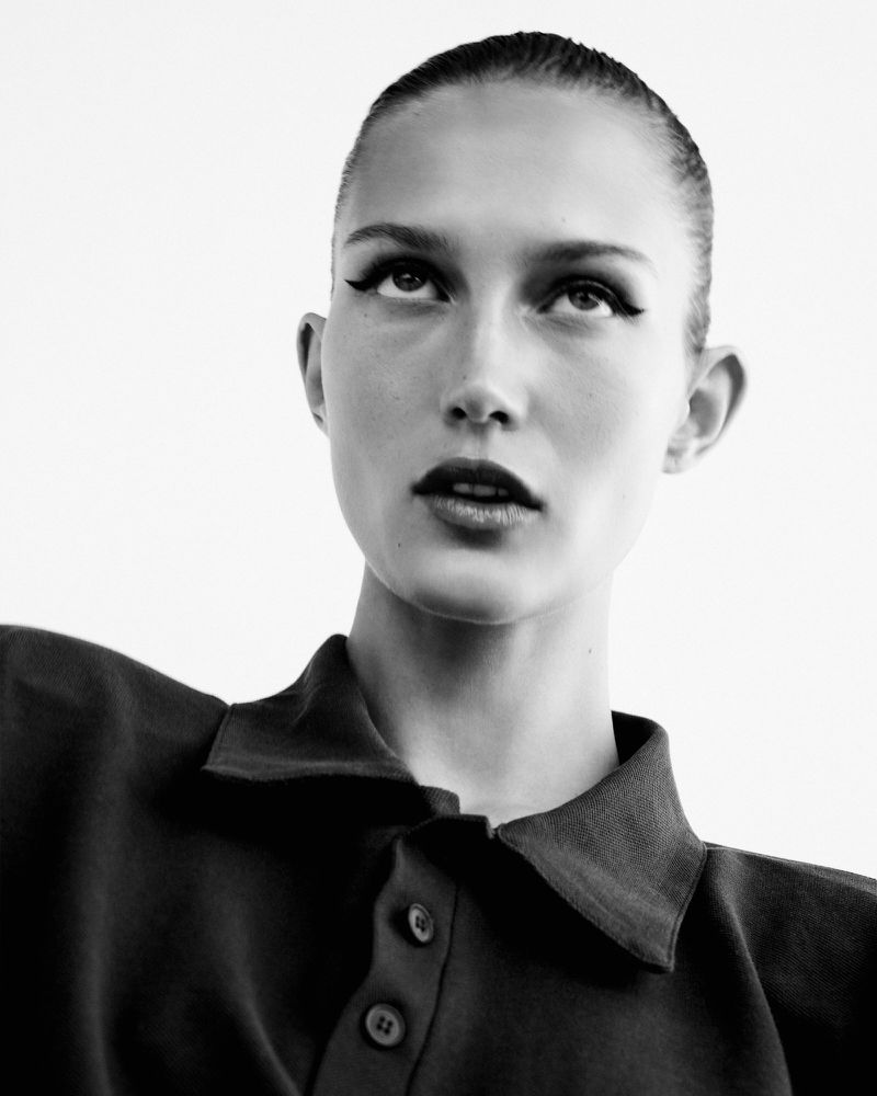 Black and white close-up of a stylish woman looking up, featuring dramatic winged eyeliner, a collared shirt, and a neutral background.