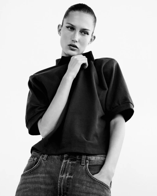 Black and white portrait of a young adult woman in a dark top and blue jeans. She is posed in front of a light background.