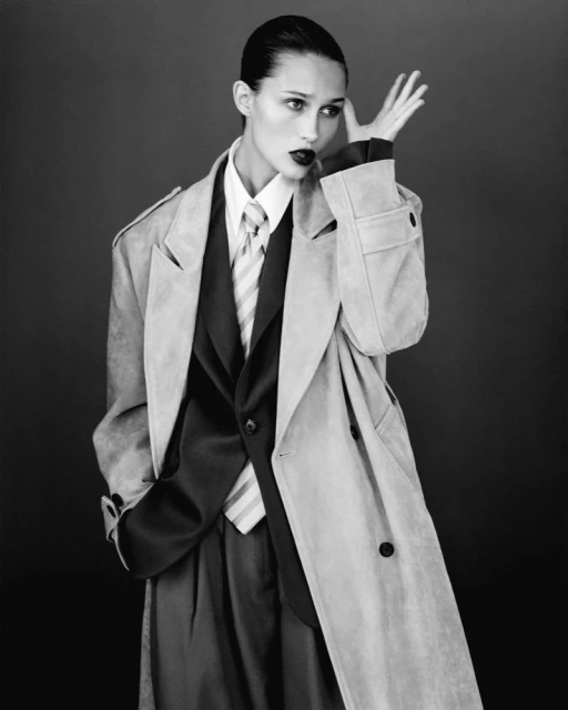 Portrait of a woman in a suit and trench coat with dark lipstick against a dark backdrop. She has slicked-back hair and is posing.