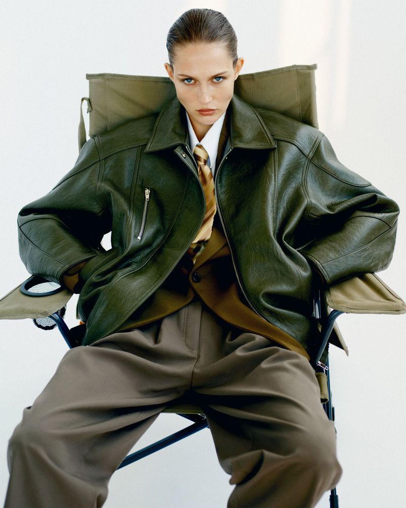 Woman in olive leather jacket sits in director's chair against a white backdrop, wearing a white shirt, tie, brown pants and blazer.