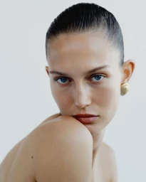 Close up portrait of model Vika Evseeva with slicked back hair and gold earrings against a white background