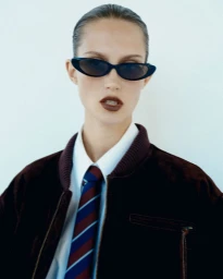 Model Vika Evseeva wearing sunglasses, a white shirt, a striped tie, and a dark brown bomber jacket against a plain background.