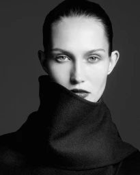Black and white portrait of model Vika Evseeva wearing an oversized black high-collar garment partially covering her face.