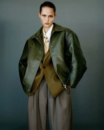 Model Vika Evseeva wearing an oversized olive leather jacket, tailored blazer, tie, and wide trousers against a grey backdrop.
