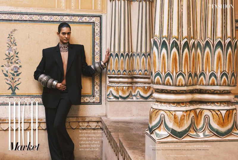 Fashion editorial shot of an adult female in a black suit and silver bracelets leaning against a patterned wall inside a building.