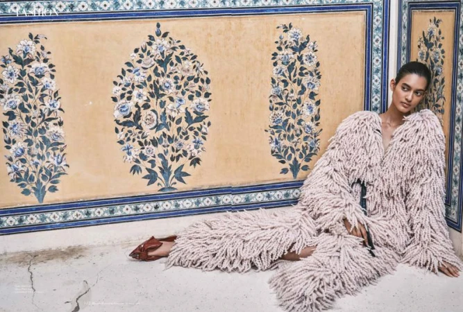 Fashion shot of a woman in a shaggy pink coat sitting against a painted wall, accented by blue floral patterns, creating a unique artistic scene.