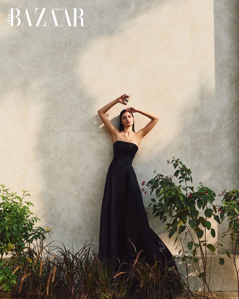 Fashion editorial shot of a woman in a black strapless dress standing in front of a marble wall with greenery, holding a perfume bottle.