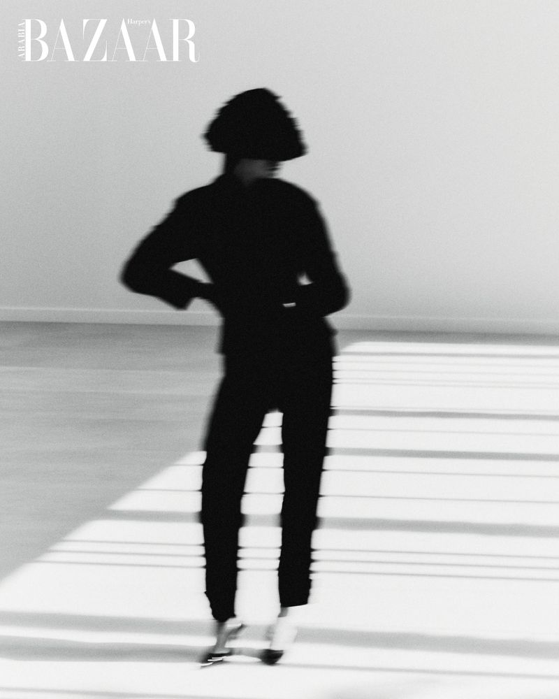 Fashion editorial shot of a silhouetted figure in black pants, jacket, and headdress, with silver shoes on a white studio backdrop.