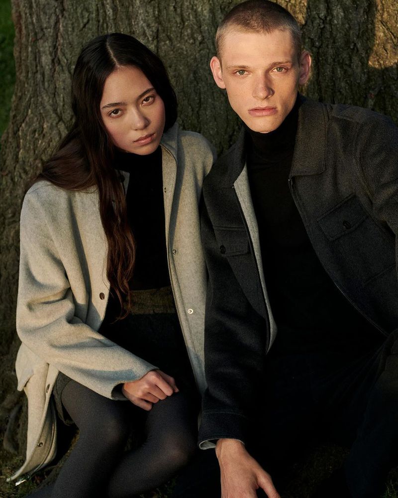 A stylish couple poses in fashionable outerwear near a tree. The outdoor setting and neutral colors create a natural, elegant scene.