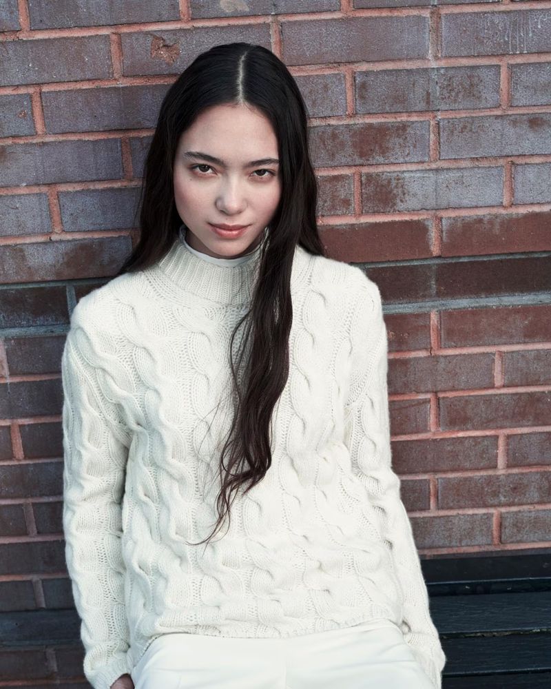 Portrait of a young adult woman with long dark hair wearing an off-white cable knit sweater, leaning against a red brick wall outdoors.