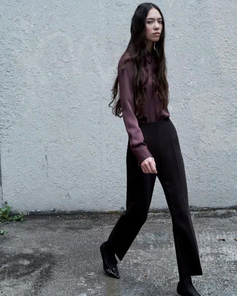 A woman walks in an outdoor setting, wearing a long-sleeve top and pants. She has long dark hair and a neutral expression.