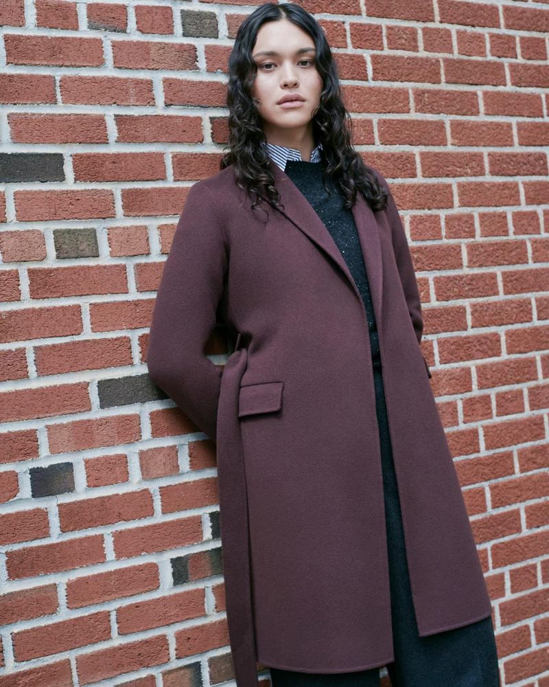 Portrait of a woman with long dark hair wearing a dark-red overcoat, posing against a brick wall backdrop in an outdoor setting.