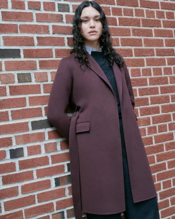 Portrait of a woman with long dark hair wearing a dark-red overcoat, posing against a brick wall backdrop in an outdoor setting.