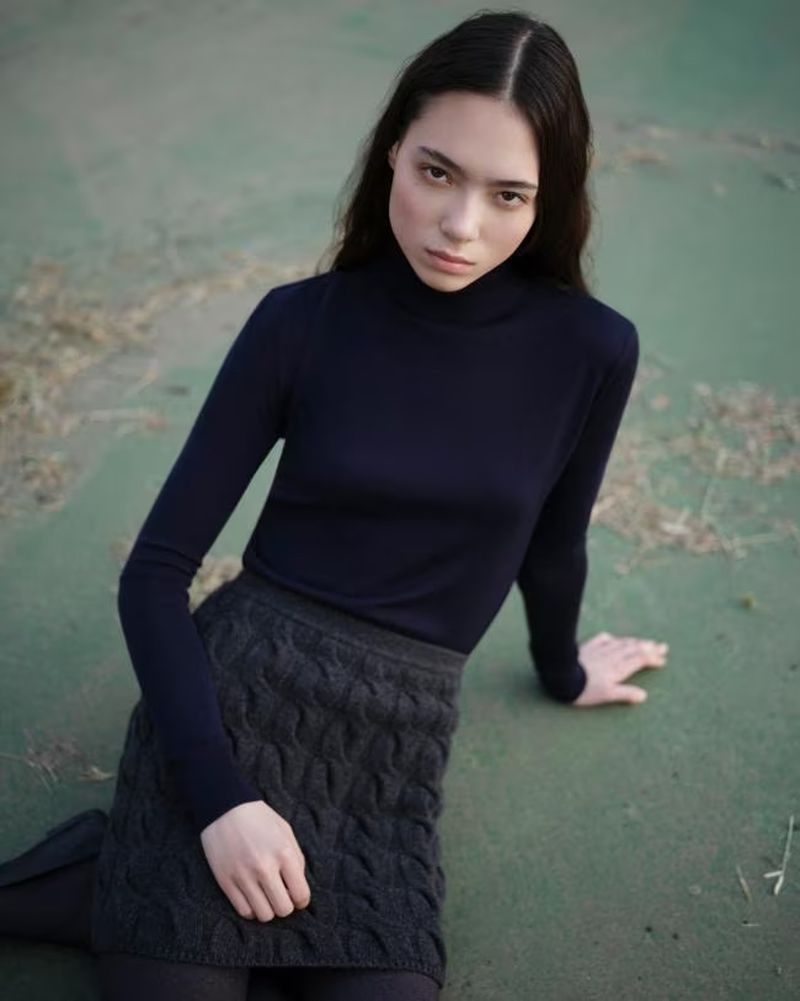 A woman with long dark hair poses in a navy turtleneck and grey knitted skirt outdoors on a muted green surface in a medium shot.