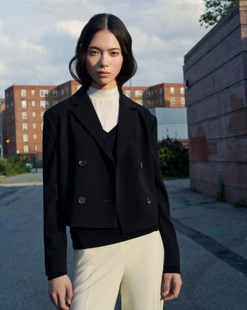 A portrait of a woman in a black blazer and cream trousers standing in an alley with brick buildings in the background.