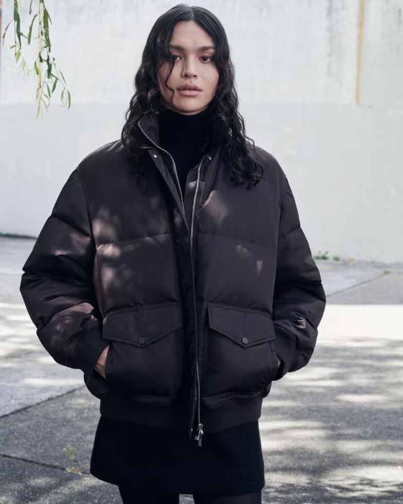 A young adult female stands outdoors wearing a brown puffer jacket, black turtleneck, and skirt, with a blurred background of trees and concrete.