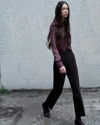 Model with long dark hair wearing a mauve silk blouse and black trousers walking past a textured grey concrete wall.