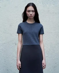 Model wearing a short-sleeved dark top and long skirt against a textured pale grey concrete wall.