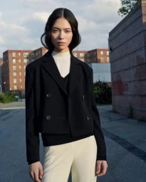 Model in a black double-breasted blazer and white trousers posing outdoors against an urban brick building backdrop.
