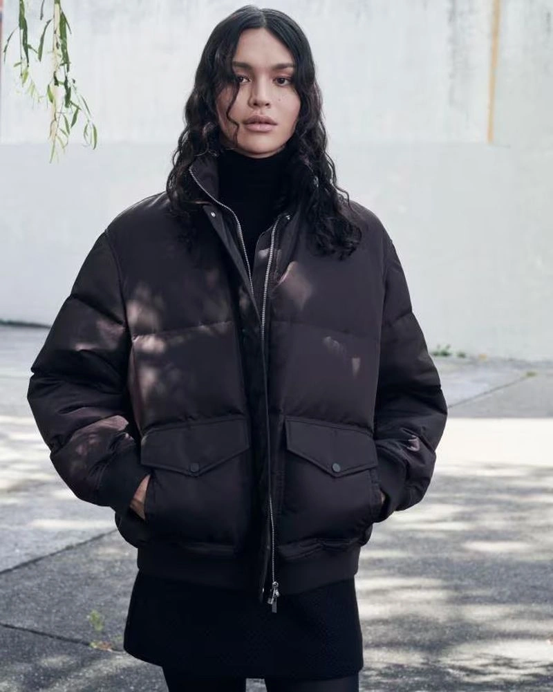 Model wearing a dark puffer jacket over a black turtleneck standing outdoors against a pale wall.