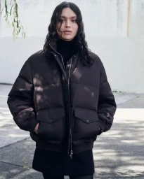 Model wearing a dark puffer jacket over a black turtleneck standing outdoors against a pale wall.