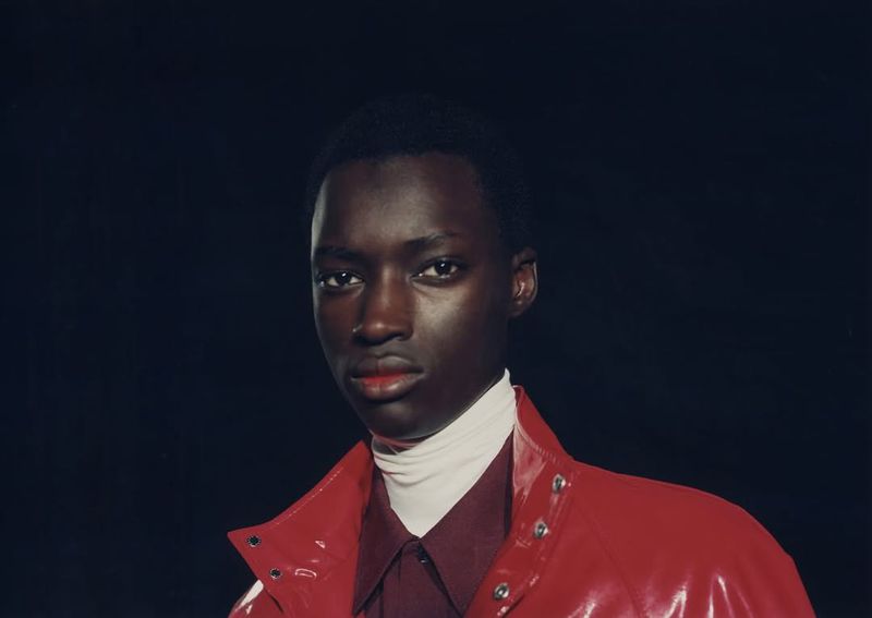 Close-up portrait of a stylish adult male with dark skin wearing a red jacket, white turtleneck, and a collared shirt against a dark backdrop.