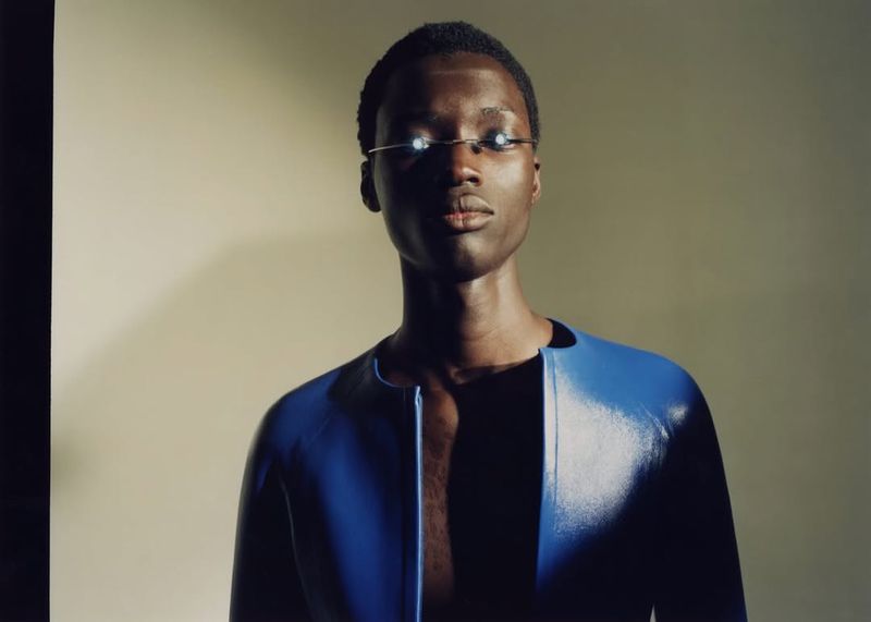 Portrait of a dark-skinned adult male wearing blue jacket with reflective glasses in a studio setting with soft, diffused lighting.