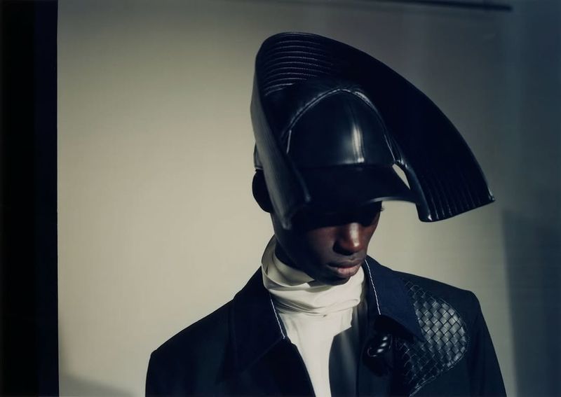 A Black male model wearing a sculptural curved black leather headpiece and dark jacket against a neutral studio backdrop.