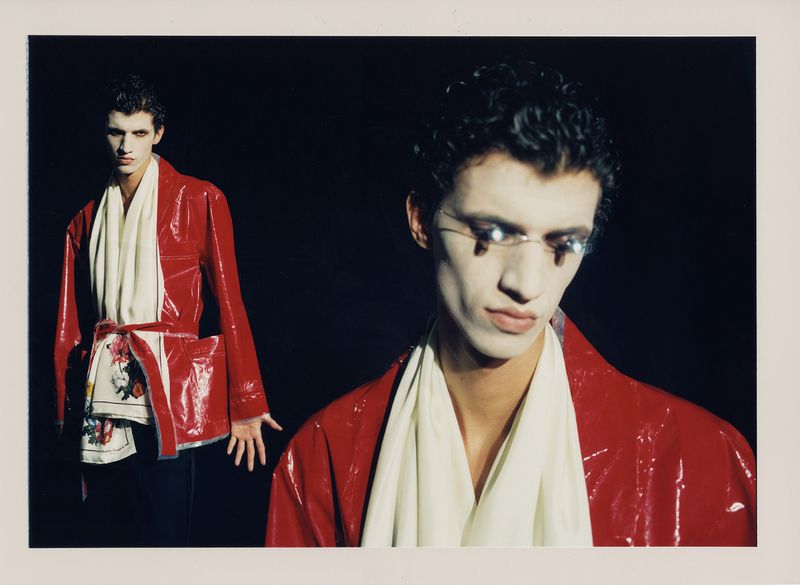 Fashionable man with white face paint and red jacket poses against a black background in an editorial portrait. Modern take on clown makeup.