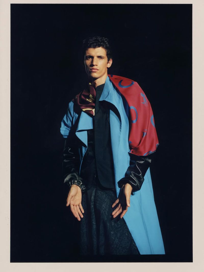 A man poses in a fashion shot against a black backdrop, wearing a light blue jacket, black top, and patterned grey trousers.