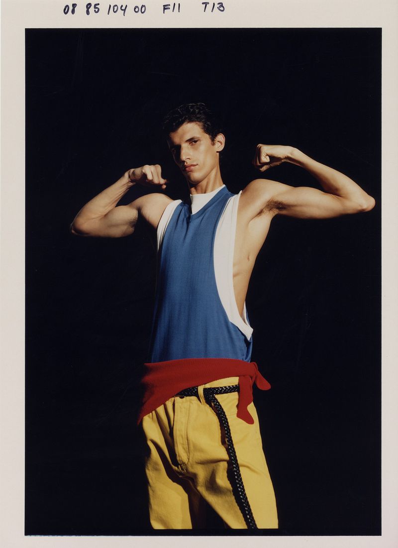 Portrait of a man with dark hair in a blue and white tank top, yellow pants, and a red sash tied around his waist, flexing his arms.