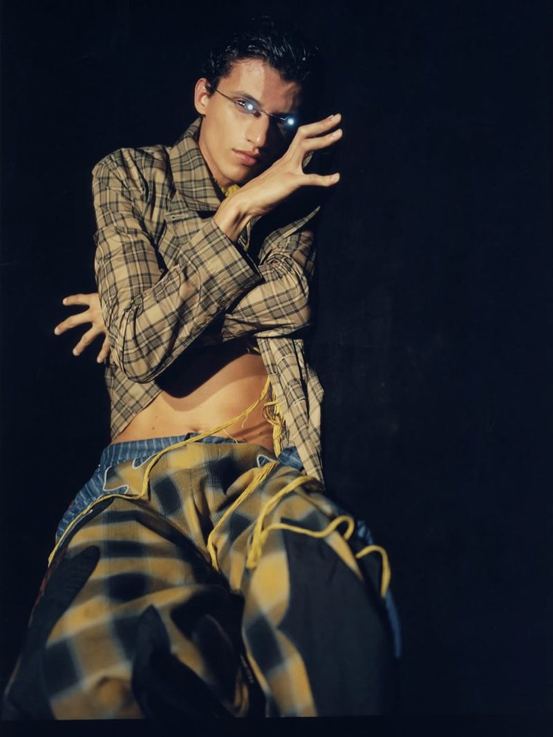 Fashion portrait of an adult male wearing a plaid jacket, trousers, and glasses, posing indoors against a black background with hands raised.