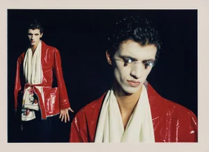 Two male models with white face makeup wearing red glossy jackets and white scarves against a black background