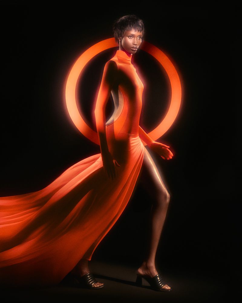 Fashion shot of a Black woman in an orange dress walking against a black background with an orange ring light behind her.
