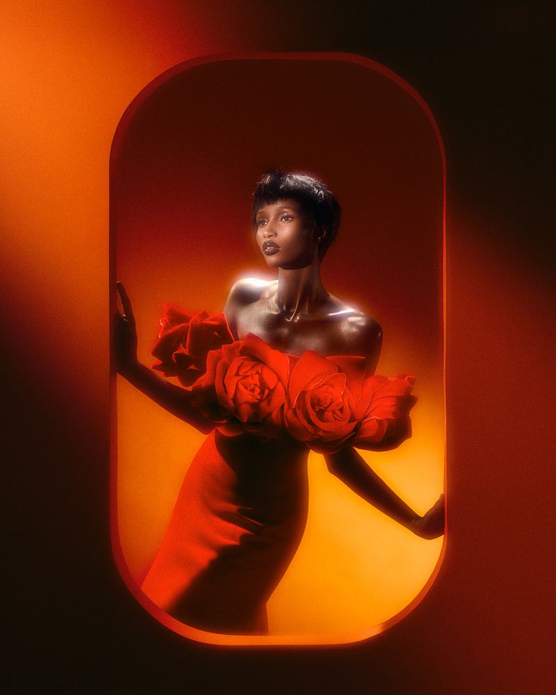 Elegant Black woman in red gown with rose detail, posing in an orange-lit arched frame, captured in a studio setting.