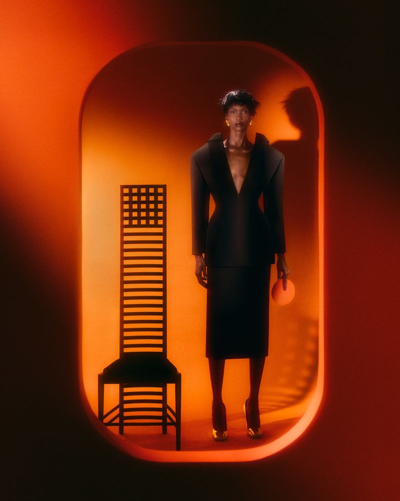 Woman in a black suit standing near a high-backed chair inside an orange tunnel, creating a high-fashion studio portrait.