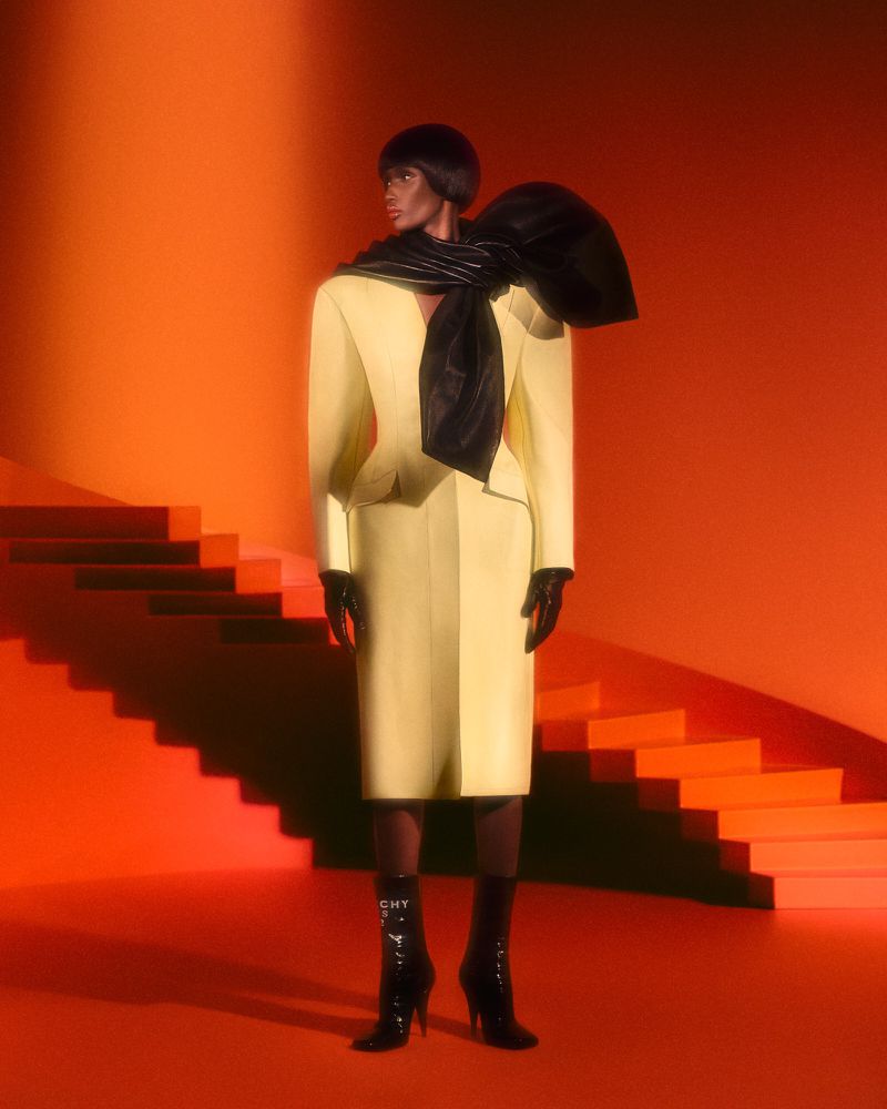 A black female model in a yellow suit and black boots poses indoors against an orange staircase and background for a fashion editorial.