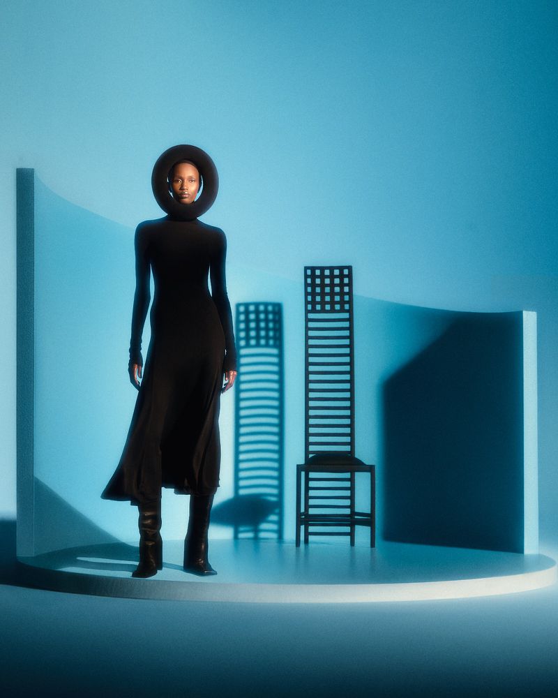 A woman in a black dress with a circular headpiece stands in a photo studio with geometric shapes and cool blue lighting.