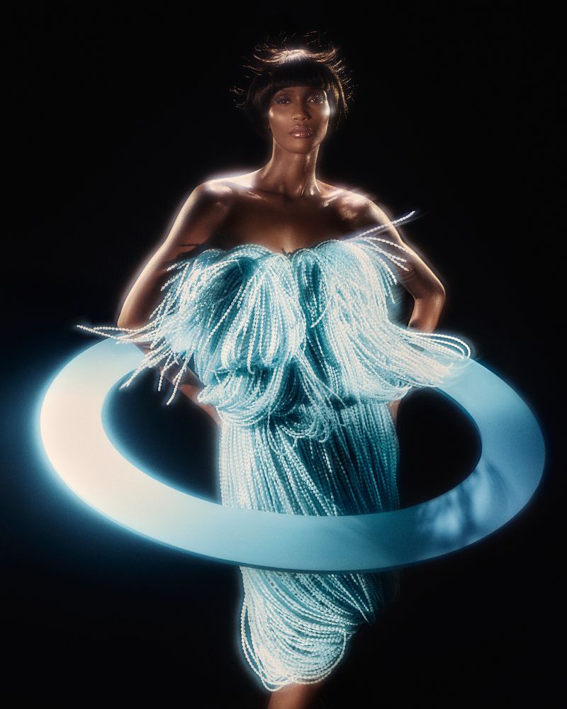Adult female model in a strapless light-blue dress with fringes and a blue hoop around the waist posing in a dark photo studio.