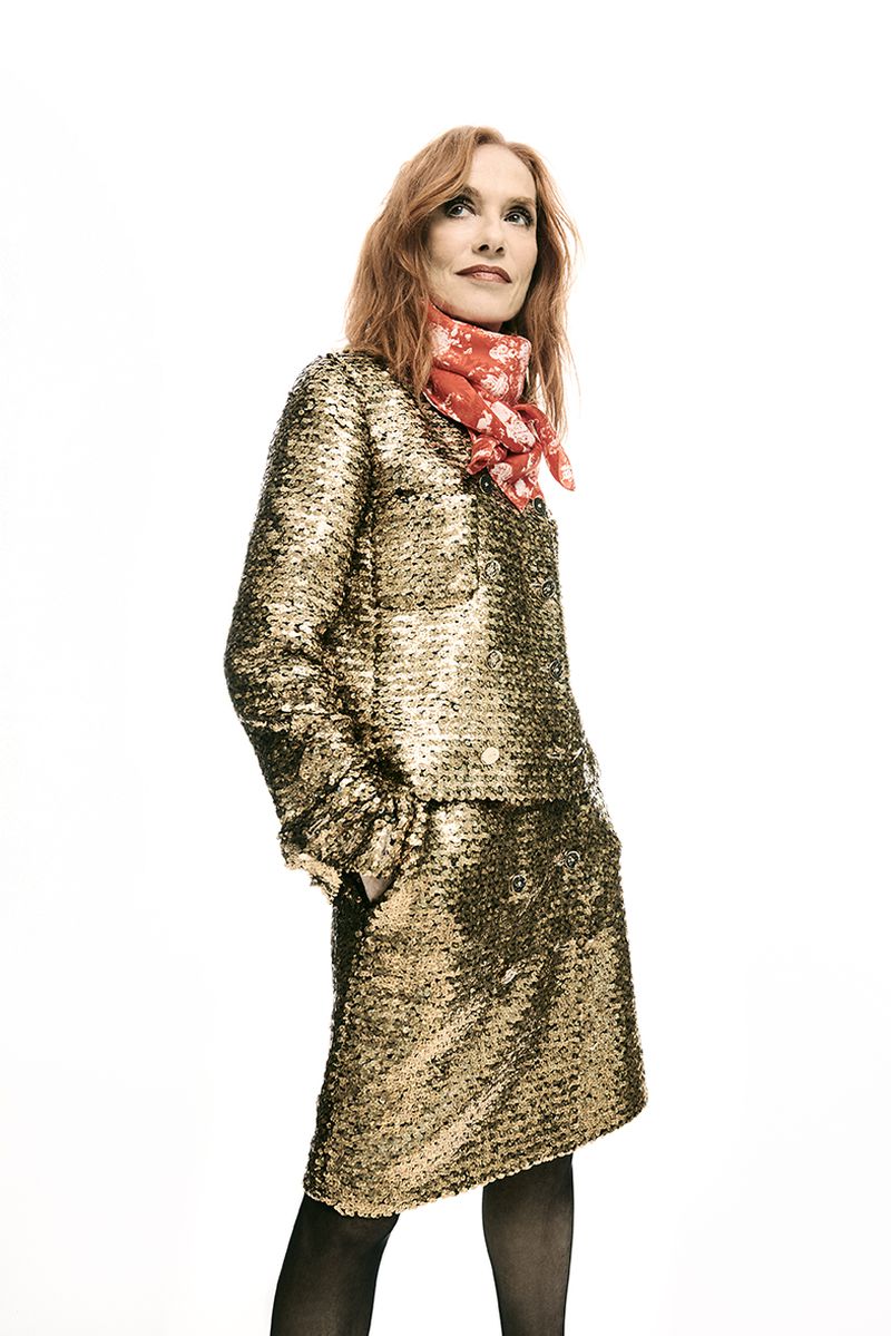 Woman with red hair wearing a gold sequined skirt suit and a floral scarf stands in a bright, neutral studio, looking up.