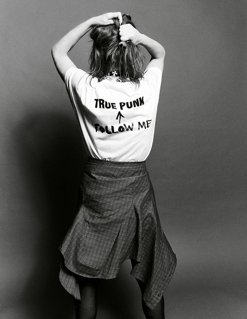 Woman viewed from behind wearing a white graphic t-shirt with True Punk Follow Me print and a draped skirt against a grey background.