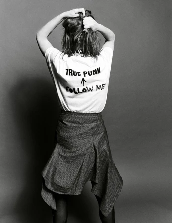 Woman viewed from behind wearing a white graphic t-shirt with True Punk Follow Me print and a draped skirt against a grey background.