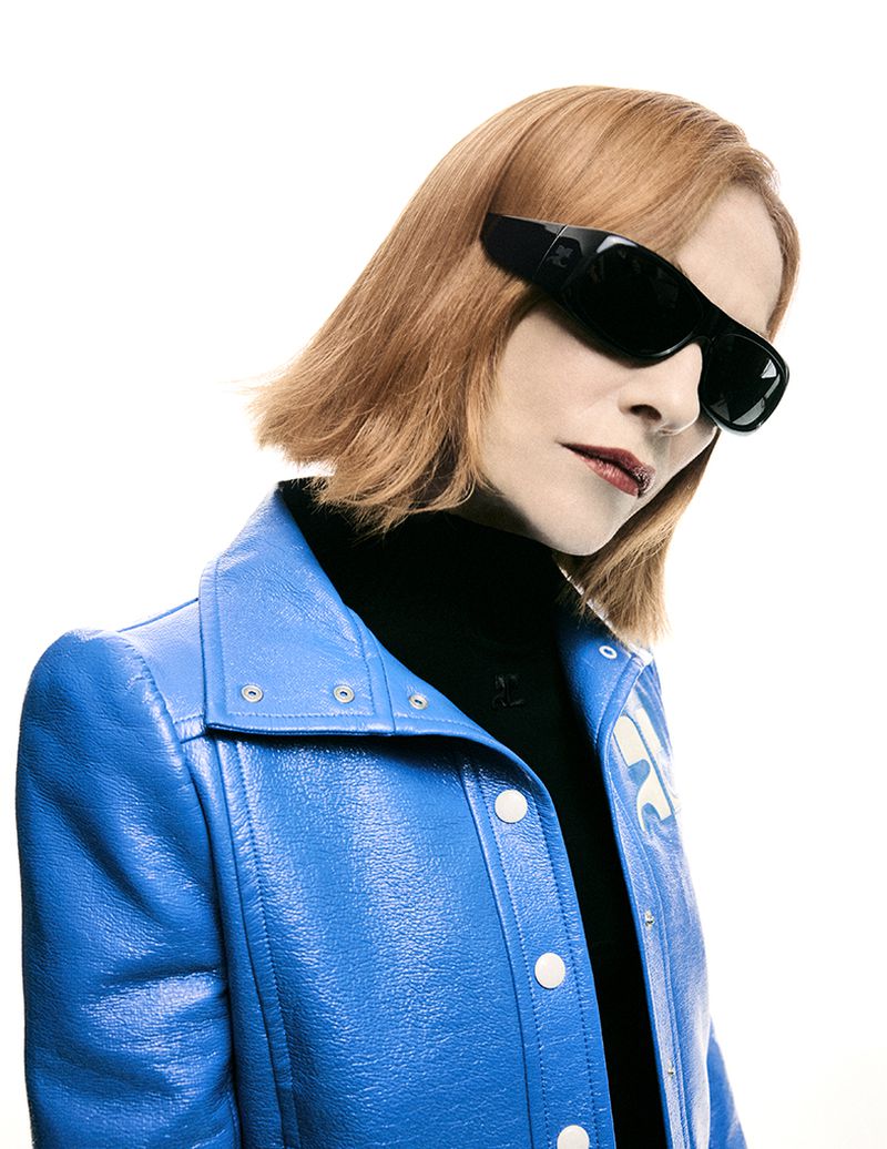 A red-haired person in a glossy blue jacket and black turtleneck, wearing oversized black sunglasses, poses against a stark white backdrop.