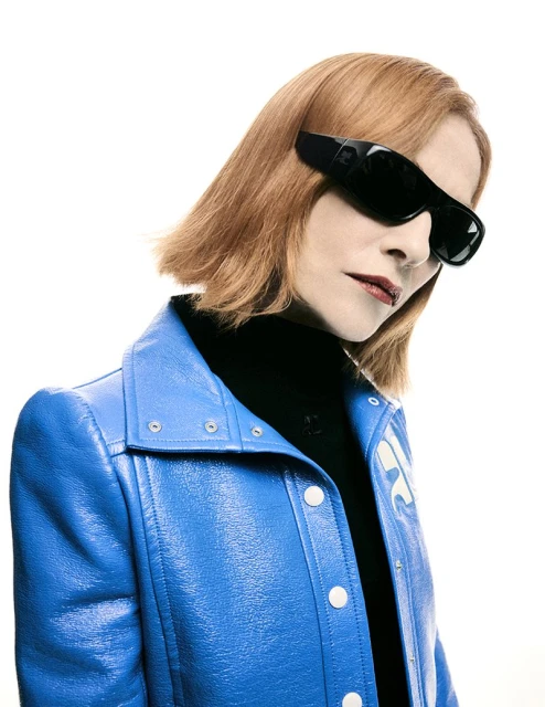 A red-haired person in a glossy blue jacket and black turtleneck, wearing oversized black sunglasses, poses against a stark white backdrop.