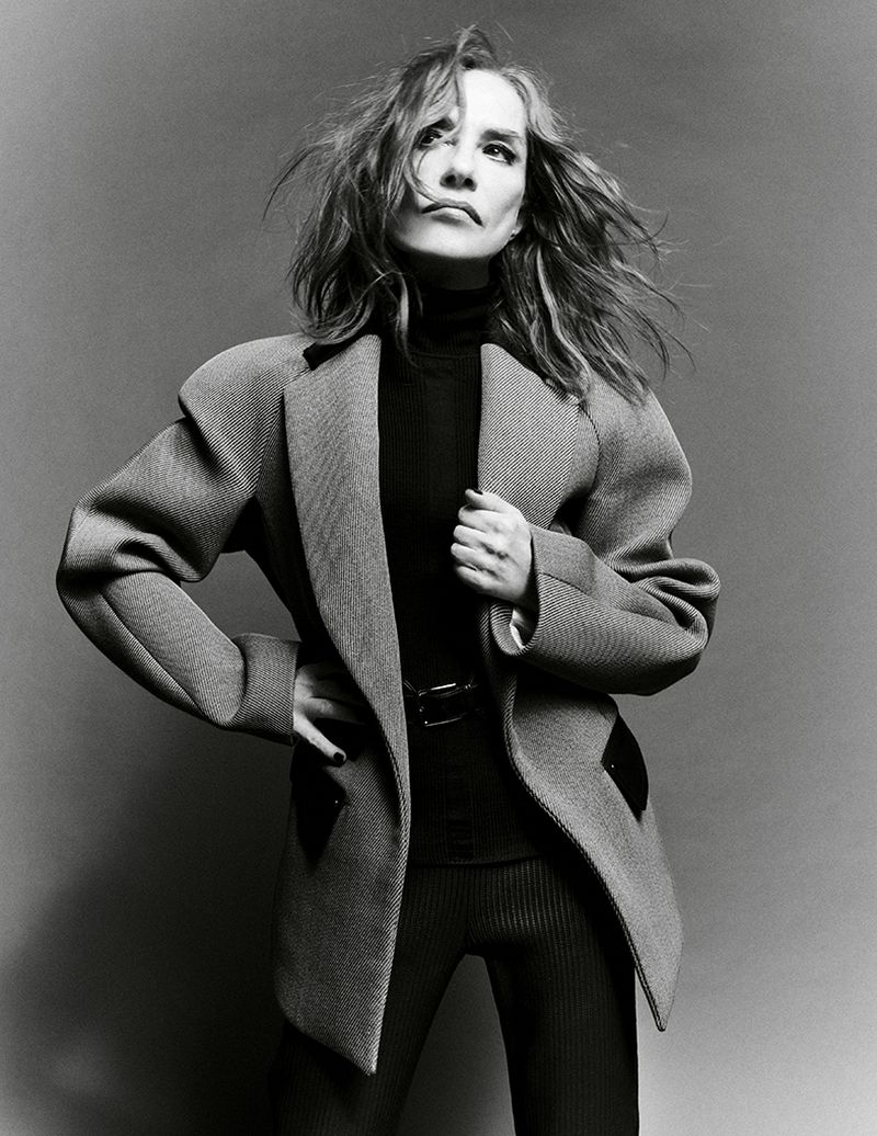 Monochrome studio shot of an adult female in a blazer, turtleneck, and leggings, striking a confident pose against a neutral backdrop.