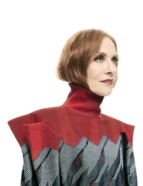 Portrait of a redhead woman in a high-necked grey and red zigzag-patterned blouse, looking to the right against a white background.